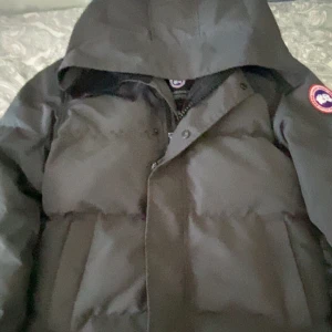 Svart dunjacka Canada Goose XS - Svart dunjacka från Canada Goose i storlek XS. Jackan har huva, klassisk logga på ärmen och dragkedja med knappslå framtill. Fylld med dun och har en puffad look som är både varm och stilren. Perfekt för kalla vinterdagar.  Pris kan diskuteras