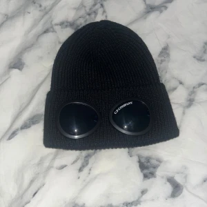 Svart mössa från C.P. Company - Svart ribbstickad mössa från C.P. Company med ikoniska goggle-linser framtill. Tillverkad i mjuk ull och har en klassisk uppvikt kant. Perfekt accessoar för att sticka ut och hålla värmen under kyliga dagar. Knappt använd, pris kan diskuteras.