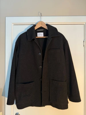 Ull Kappa - Kappa i mörkgrå ull från American Vintage med klassisk krage, tre stora framfickor och knäppning med knappar. Långärmad och rak passform, perfekt under höst och vinter. Storlek S för lite längre passform, storlek M för en lite kortare ”vanlig” passform!