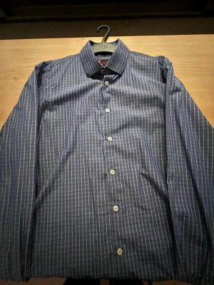 Blårutig skjorta med knappar - Snygg blårutig skjorta med vita knappar och klassisk krage. Skjortan har lång ärm och är tillverkad i ett mjukt bomullstyg som känns skönt mot huden. Perfekt för dig som gillar stilrena mönster och enkel design.