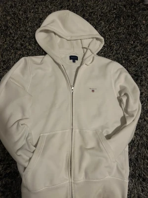 Vit zip hoodie från GANT - Säljer en stilren vit hoodie från GANT i storlek M. Hoodien har dragkedja framtill, två fickor och en broderad GANT-logga på bröstet. Tillverkad i mjukt bomullsmaterial och har en klassisk huva. Perfekt för en clean och avslappnad look.