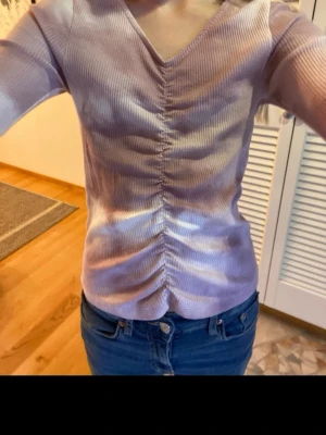 Ljuslila ribbad topp med v-ringning - Säljer en ljuslila ribbad topp med v-ringning och rynkade detaljer framtill. Toppen har lång ärm och är figurnära i modellen. Perfekt att styla med jeans eller kjol för en trendig look.