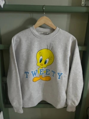 Grå Tweety sweatshirt Looney Tunes - Säljer en grå sweatshirt med stor Tweety-tryck framtill och blå text. Tröjan har rund halsringning och ribbade muddar vid ärmslut och nederkant. Perfekt för dig som gillar retro och tecknade figurer.