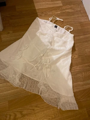 Vit omlottklänning från SHEIN - Söt vit omlottklänning från SHEIN med tunna axelband och volangdetaljer. Klänningen har snörning framtill och transparenta partier med prickigt mönster längst ner. Perfekt för sommarens alla tillfällen.