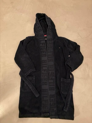 Svart morgonrock Tommy Hilfiger - Mysig svart morgonrock från Tommy Hilfiger med huva, två stora fickor och bälte i midjan. Tillverkad i mjuk frotté och har logotypen diskret broderad samt tryckt längs kanten. Perfekt för chilliga morgnar eller kvällar hemma. Large ungdom, passar för folk runt 160