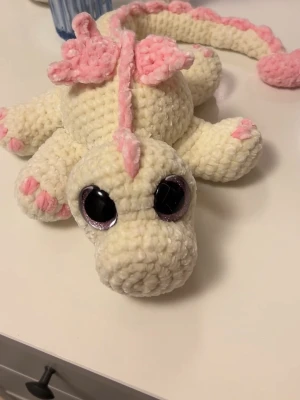 Vit drake med ljusrosa detaljer🤍💕 - Denna söta drake letar efter sitt nya framtida hem🐉🤍 Denna är virkad med kärlek, jag älskar att virka🤍🩷 Den är virkad av mjukt garn från Rusta, hårda ögon som inte går att ta av. Den är cirka 56cm lång om man sträcker ut svansen🐉
