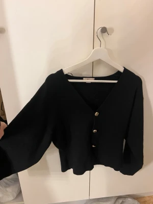 Svart kofta med guldknappar - Stilren svart kofta med v-ringning och tre dekorativa guldknappar framtill. Koftan har en något kortare och boxig passform samt långa, lite vidare ärmar. Perfekt att slänga över en topp för en trendig look.