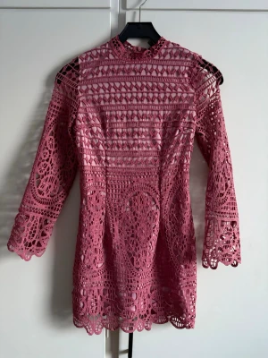 Rosa spetsklänning med trekvartsärm - Säljer en superfin rosa kortklänning i spets med trekvartsärm och hög hals. Klänningen har ett detaljerat spetsmönster och är figurnära med öppen axeldetalj. Helt ny med lapp kvar.