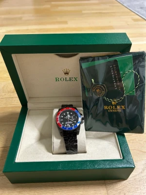 klocka svart/blå/röd bezel - Säljer en snygg klocka med svart länkarmband och en bezel i blått och rött. Klockan har svart urtavla med vita detaljer och levereras i originalbox med certifikat. Perfekt för dig som vill ha en ikonisk och stilren accessoar.