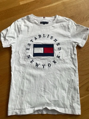 Vit t-shirt från Tommy Hilfiger - Vit t-shirt från Tommy Hilfiger med det klassiska logotrycket i blått, vitt och rött på bröstet samt texten 'Established New York' runt loggan. T-shirten har rund hals och korta ärmar. Tillverkad i mjuk bomull för en skön känsla.