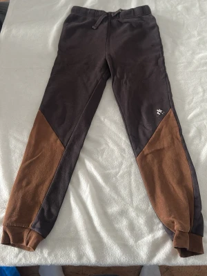 Bruna och mörkbruna mjukisbyxor - Snygga mjukisbyxor i mörkbrunt med bruna paneler på benen. Byxorna har resår i midjan med snörning och muddar vid bensluten. Liten vit logga på ena benet. Perfekta för chill eller träning.