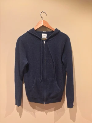 Mörkblå hoodie med dragkedja - Mysig mörkblå hoodie med dragkedja framtill och två fickor. Tillverkad i ett mjukt, stickat material och har en klassisk huva. 100% kashmere som är i nyskick. Storlek S.