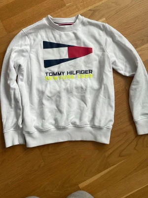 Vit sweatshirt Tommy Hilfiger - Vit sweatshirt från Tommy Hilfiger med stort tryck och broderad logga framtill. Tröjan har rund halsringning, ribbade muddar och detaljer i marinblått, rött och gult. Texten 'NEW YORK 1985' i neonfärg ger en sportig vibe.
