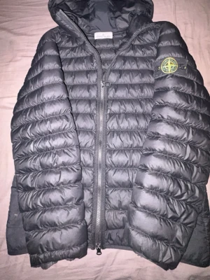 Svart pufferjacka från Stone Island - Säljer en svart pufferjacka från Stone Island med huva och klassisk patch på ärmen. Väldigt bra skick och använd några fåtal gånger i vinter.Jackan har quiltad design, dragkedja framtill och är tillverkad i lätt polyester. Perfekt för kalla dagar och riktigt snygg streetstil. Priset är diskuterbart eftersom den är äkta men lite använd 