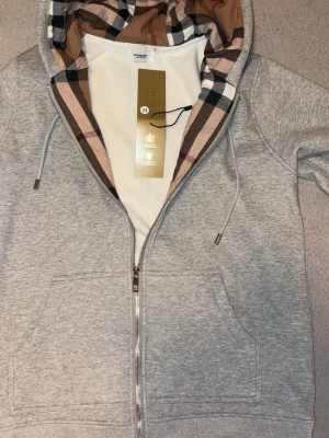 Grå hoodie från Burberry med huva - Snygg grå zip-hoodie från Burberry med klassiskt rutigt mönster i huvan. Tillverkad i mjuk bomull och har dragkedja framtill samt ribbade muddar. Perfekt för en avslappnad och trendig look. Ikonisk Burberry-detalj i innerfodret på huvan. Hoodien i bilden syns tydligt utan några repor eller skador om ni vill ha mer bilder det är bara att säga till annat har jag ingen retur eller öppet köp