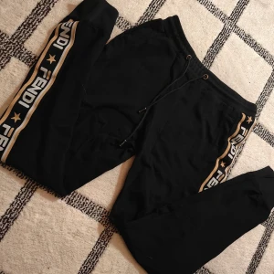Svarta mjukisbyxor från Fendi - Fila x fendi 2018 collab. Svarta mjukisbyxor från Fendi med guldfärgade och vita detaljer längs sidorna. Byxorna har snörning i midjan och Fendi-logga med stjärnor på banden. Perfekta för en chill och trendig look.