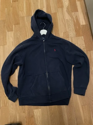 Mörkblå hoodie från Ralph Lauren - Mörkblå hoodie med dragkedja från Ralph Lauren. Klassisk design med huva, kängurufickor och den ikoniska röda loggan broderad på bröstet. Tillverkad i mjukt bomullsmaterial, perfekt för en avslappnad och stilren look.