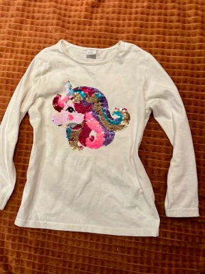 Vit långärmad topp med paljett-Enhörning - Vit långärmad topp med en stor färgglad enhörning i paljetter på framsidan. Enhörningen har inslag av rosa, blått, guld och lila. Rund halsringning och mjukt bomullsmaterial. Perfekt för dig som gillar magiska detaljer och vill sticka ut.