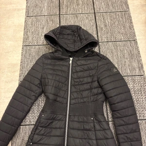 Svart pufferjacka från Moose Knuckles - Svart pufferjacka från Moose Knuckles med huva, markerad midja och snygga quiltade detaljer. Jackan har dragkedja framtill och är tillverkad i polyester med återvunna material. Perfekt för kalla dagar med sin vind- och vattenavvisande funktion. Jackan är inte använd mycket alls köptes ny på miinto 