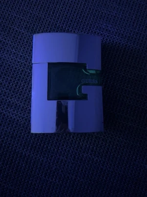 Guess Man Eau de Toilette - Guess Man Eau de Toilette med en modern, stilren flaska i silverfärgad metall och grönt glas. Flaskan har en unik, kantig form och Guess-loggan syns tydligt på framsidan. Perfekt för dig som gillar maskulina och fräscha dofter.