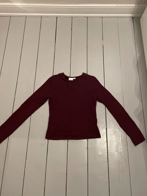 Vinröd långärmad topp med v-ringning - Vinröd långärmad topp med v-ringning och diskreta spetsdetaljer vid halsen. Tunn och mjuk kvalitet som sitter snyggt på kroppen. 