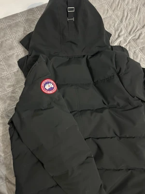 Svart dunjacka från Canada Goose - Säljer en svart dunjacka från Canada Goose med klassisk patch på ärmen och stor huva med spännen. Jackan har dragkedja och tryckknappar framtill samt två sidofickor. Perfekt för kalla vinterdagar och riktigt snygg streetstil. Kvitto tillkommer 