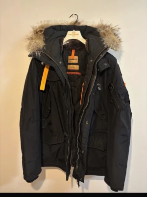 Svart Parajumper !!ÄKTA!! - Säljer en svart Parajumper med avtagbar huva och päls, flera fickor och gul detaljrem. Jackan har dragkedja och knappar framtill samt lång ärm. Perfekt för kalla dagar och riktigt snygg streetstil.