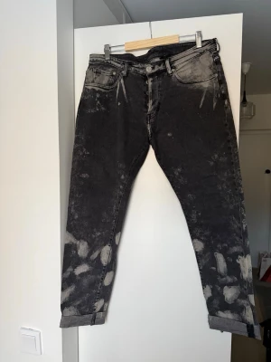 Svarta jeans med blekta detaljer - Unika svarta jeans med coola blekta fläckar och tvättad look. Jeansen har klassisk femficksmodell, normal passform och uppvikta benslut. Perfekta för dig som vill sticka ut med en edgy stil. Materialet är tålig denim i bomull.