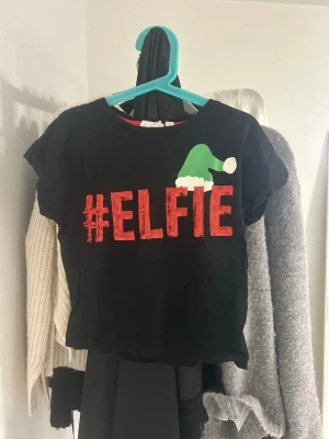 Svart croppad jul-tshirt #ELFIE - Svart croppad t-shirt med röd texten #ELFIE framtill och en grön tomteluva som detalj. Perfekt för dig som vill ha en rolig och festlig look till julen. T-shirten har korta ärmar och är gjord i mjuk bomull.