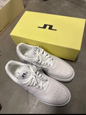 Vita sneakers från J.Lindeberg - Stilrena vita sneakers från J.Lindeberg med klassisk låg profil och perforerad tå. Skorna har snörning, rund tå och är tillverkade i slätt syntetmaterial med diskret logga på sidan och plös. Perfekta för en clean och modern look.