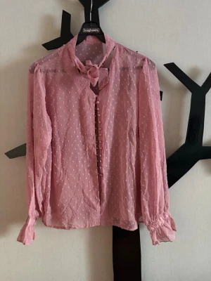 Rosa prickig blus med knytband - Söt rosa blus med små prickar, knytband i halsen och dekorativa knappar framtill. Blusen är i ett tunt, transparent tyg och har långa ärmar med resår vid ärmslut. 