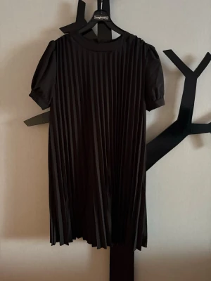 Svart plisserad kortklänning - Svart klänning msd puffiga korta ärmar. Klänningen har rund halsringning och är tillverkad i ett mjukt, lätt material som ger ett snyggt fall. Knapp på ärmen. Är tyg under, så den är rejäl.