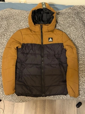 Pufferjacka från H&M - Snygg pufferjacka från H&M i brun och svart med huva och dragkedja framtill. Jackan har en quiltad look och en patch på bröstet. Perfekt för kalla dagar och riktigt skön att bära.