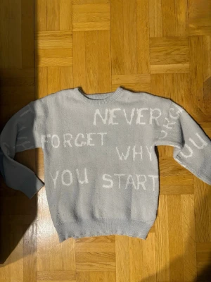 Ljusgrå stickad tröja med text - Ljusgrå stickad tröja med vit text över hela framsidan som säger 'NEVER FORGET WHY YOU START'. Tröjan har rund halsringning, långa ärmar och ribbstickade muddar vid ärmslut och nederkant. Perfekt för en avslappnad och trendig look.