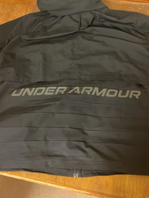 Svart vindjacka från Under Armour - Snygg svart vindjacka från Under Armour med stor logga på ryggen och dragkedja framtill. Jackan har huva och är tillverkad i lätt polyester, perfekt för blåsiga dagar. Enkel och sportig design som funkar till det mesta.