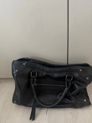 Svart handväska från Gina Tricot - Snygg svart handväska från Gina Tricot i mjukt syntetmaterial. Väskan har silverfärgade metalldetaljer, dubbla handtag och en dragkedja framtill. Perfekt storlek för att få plats med allt du behöver och passar till många olika outfits.