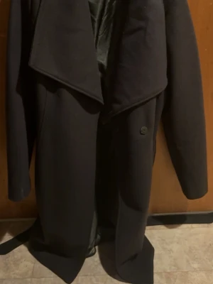 Svart kappa från VILA med bälte - Stilren svart kappa från VILA med bred krage och bälte i midjan. Kappan har lång ärm och en klassisk, elegant look som passar perfekt till höst och vinter. Tillverkad i ett mjukt material med slät yta och helfodrad insida.