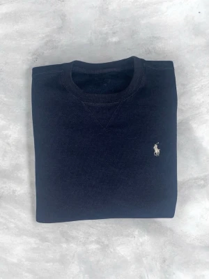 Mörkblå tröja från Ralph Lauren - Klassisk mörkblå tröja från Ralph Lauren med rund halsringning och diskret broderad logga på bröstet. Tröjan har långa ärmar och ribbade muddar vid ärmslut och nederkant. Perfekt för en stilren och enkel look.