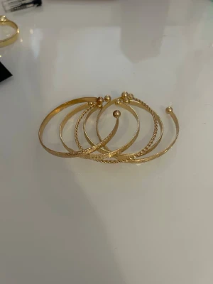 Set med fem guldiga armband - Säljer ett set med fem snygga armband i guldfärgad metall. Armbanden har olika texturer och mönster, bland annat släta, tvinnade och räfflade ytor. Alla är justerbara i modellen och avslutas med små kulor i ändarna. Perfekt för att mixa och matcha till din stil.