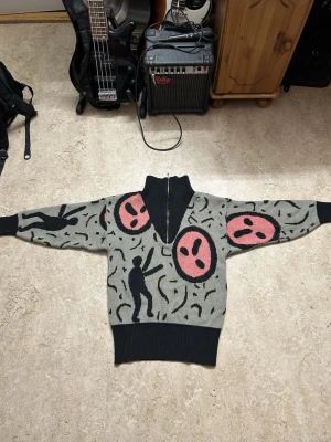Grå stickad tröja med grafiskt mönster - Unik grå stickad tröja med svarta och rosa grafiska motiv, halvzip och hög krage. Tröjan har svarta siluetter och stora rosa ansikten, samt breda svarta muddar vid ärmslut och midja. Perfekt för dig som gillar statement-plagg och kreativ design.