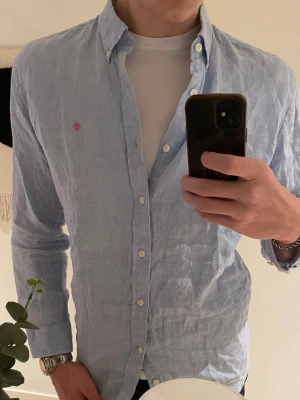 Ljusblå linneskjorta från Morris - Säljer en ljusblå skjorta från Morris i linne med klassisk button-down krage och vita knappar. Skjortan har en liten rosa broderad detalj på bröstet och en avslappnad passform. Perfekt för dig som gillar stilrena och svala plagg.