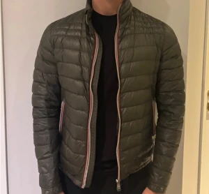 Grön Jacka Från Moncler - Snygg mörkgrön dunjacka från Moncler med quiltad design och klassisk logga på ärmen. Jackan har dragkedja framtill och randiga detaljer längs dragkedjan och fickorna. Perfekt för kalla dagar och ger en clean, modern look.