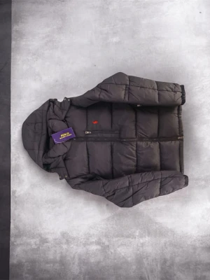 Svart pufferjacka från Polo Ralph Lauren - Snygg svart pufferjacka från Polo Ralph Lauren med huva och quiltad design. Jackan har dragkedja framtill och en liten röd logga på bröstet. Perfekt för kalla dagar och ger en modern, sportig look.