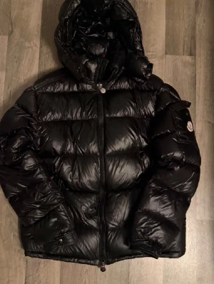 Svart dunjacka från Moncler - Snygg svart dunjacka från Moncler med glansig finish och klassisk Moncler-logga på ärmen. Jackan har huva, dragkedja framtill och quiltad design som ger en riktigt puffig look. Perfekt för kalla dagar och riktigt trendig stil.
