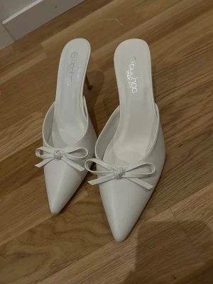 Vita pumps med rosett från Boohoo - Stilrena vita pumps från Boohoo med spetsig tå och smal klack. Skorna har en elegant rosettdetalj framtill och är tillverkade i syntetmaterial. Perfekta för att lyfta din outfit med en chic touch. Använda 1-2 ggr