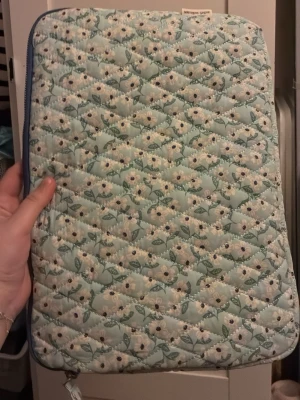 Oandvänd trendig Blommig quiltad datorväska från systrarna grene  - Funkar för hp skoldagar, Säljer en quiltad datorväska i ljusblått med blommigt mönster. Väskan har dragkedja och är vadderad för extra skydd. Perfekt för dig som vill ha en söt och unik laptopväska med romantisk vibe.