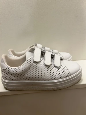 Vita sneakers med kardborre från Victoria - Säljer ett par vita sneakers från Victoria med tre kardborreband och perforerat mönster på sidorna. Skorna har platt sula och något platå. Använda 2 ggr. Har inte testat göra rent sulan men går nog att ta bort. Perfekta till våren