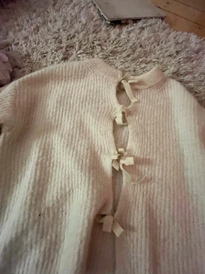 Krämvit stickad kofta med rosetter - Supersöt krämvit stickad kofta från Gina Tricot med dekorativa rosetter längs magen. Koftan har rund halsringning och är ribbstickad för en mysig känsla. Perfekt för lager-på-lager och en gullig touch till din outfit.