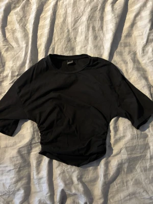 Svart croppad t-shirt med scrunch i sidorna - En svart croppad t-shirt med scrunch i sidorna. T-shirten har korta ärmar och rund halsringning. Perfekt för en trendig och avslappnad stil. Tillverkad i mjuk bomull för skön känsla.