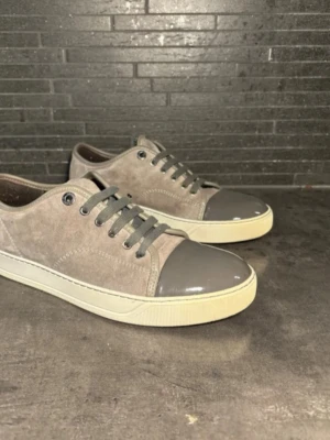 Gråa Lanvins mockasneakers med lackdetalj - Snygga Lanvins sneakers i grå mocka med matchande grå snörning och glansig lackad tå. Skorna har en vit platt sula och stilren design som passar till det mesta. Perfekta för dig som gillar en clean och modern look.